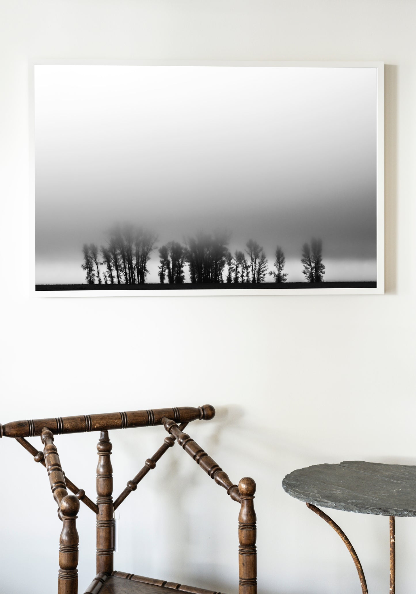 Framed black and white landscape of trees on a foggy day above a wooden chair and table.