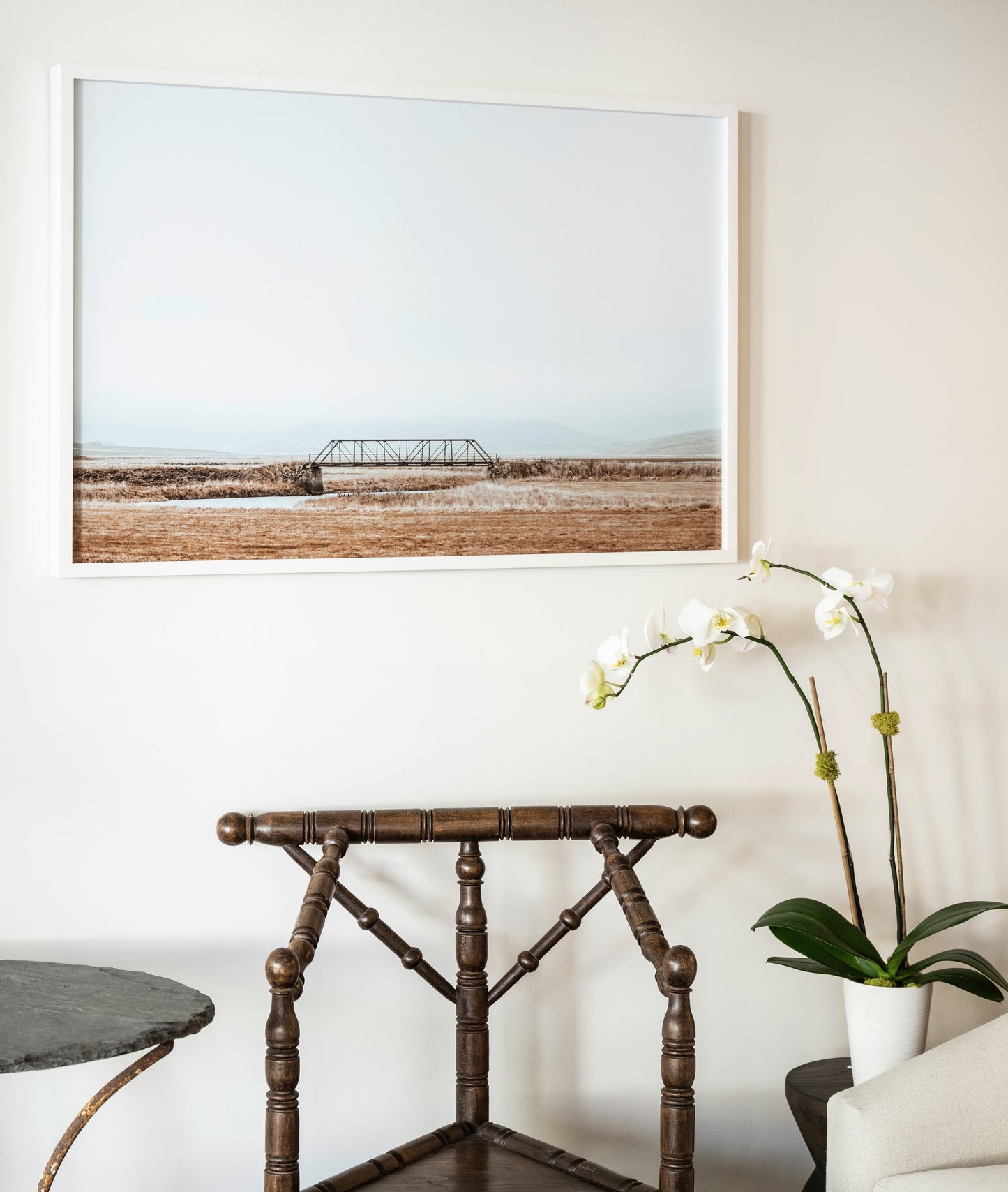Framed landscape print on a wall above a wooden chair with a plant.