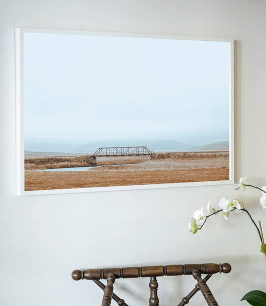 Framed artwork of a landscape with a bridge on a wall above a wooden chair.