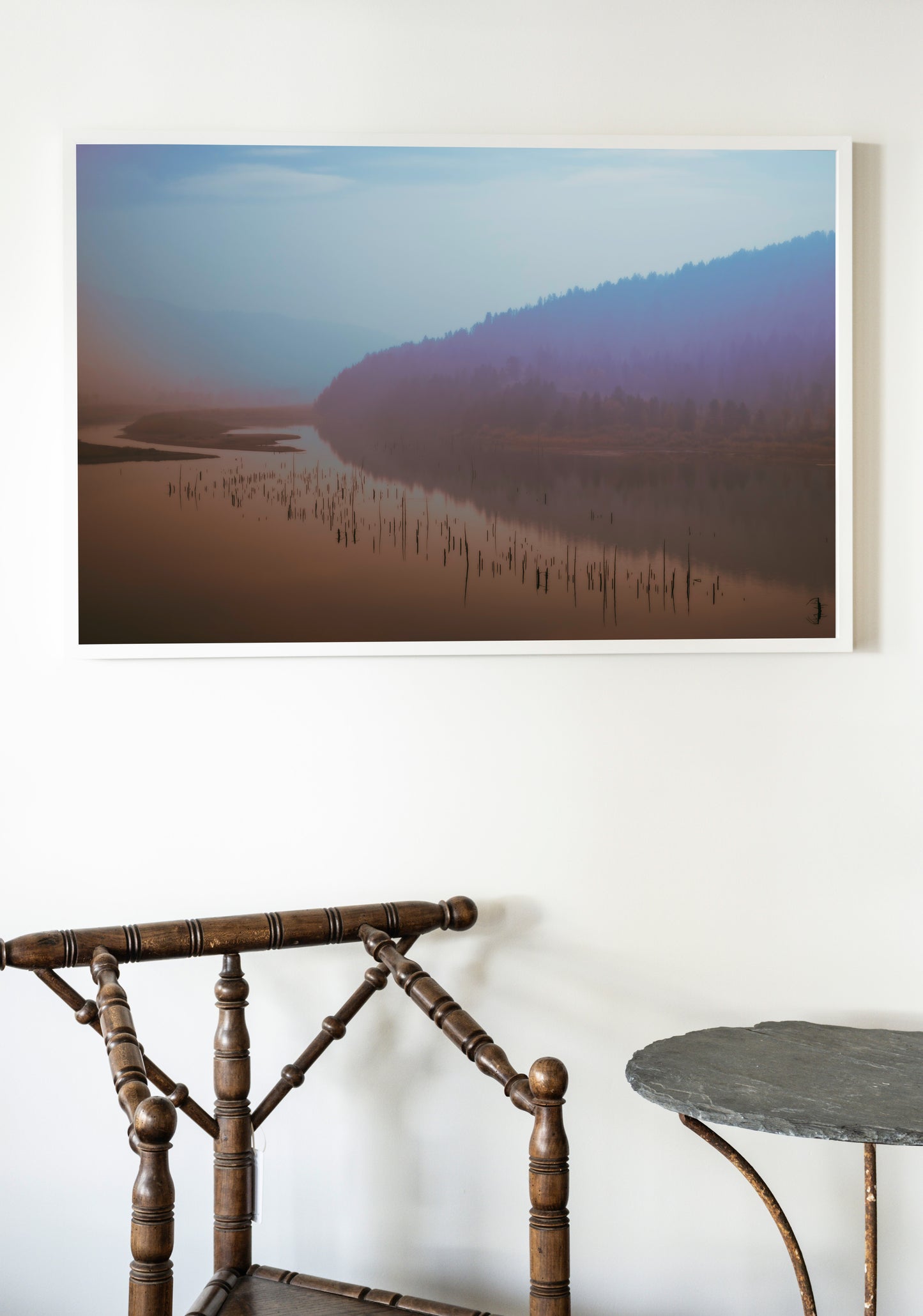 Framed photograph of a misty landscape with a lake and mountains on a white wall above a wooden chair and table.