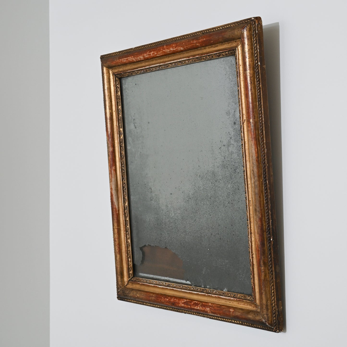 An Early 19th Century Mercury Glass Mirror with a rectangular, distressed gilt wood frame hangs on a white wall; its heavily tarnished surface shows dark spots and significant damage near the bottom edge.