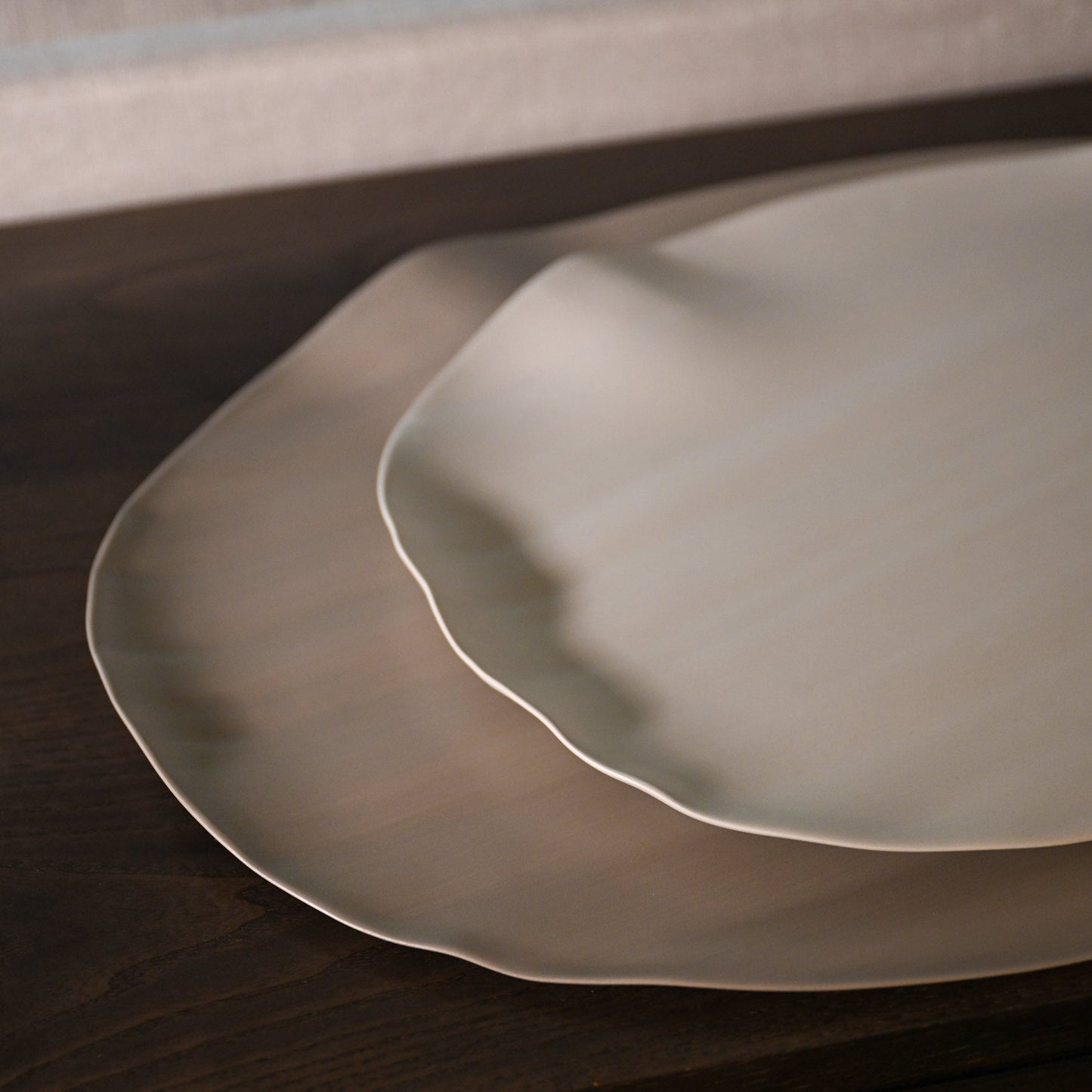 Two matte off-white ceramic Leaf Large linen plates with wavy edges stacked on a dark wooden surface
