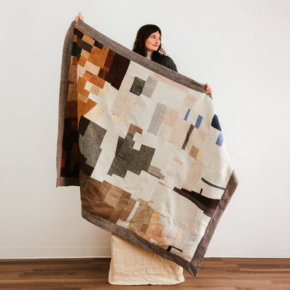 Large abstract patchwork quilt with geometric blocks in beige, brown, gray, and muted blue tones.