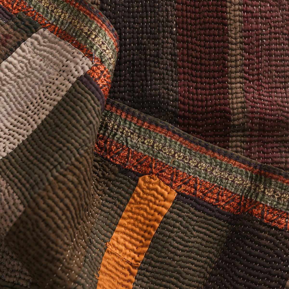 Close-up of a patchwork quilt with textured woven panels in dark brown, olive green, and rust orange.