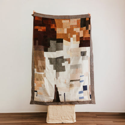 An abstract patchwork quilt with geometric blocks of brown, beige, white, gray, and muted blue.