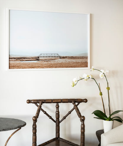 Framed landscape print on a wall above a wooden chair with a plant.
