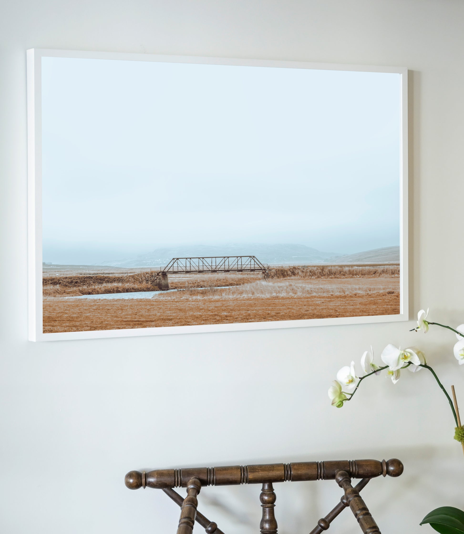 Framed artwork of a landscape with a bridge on a wall above a wooden chair.