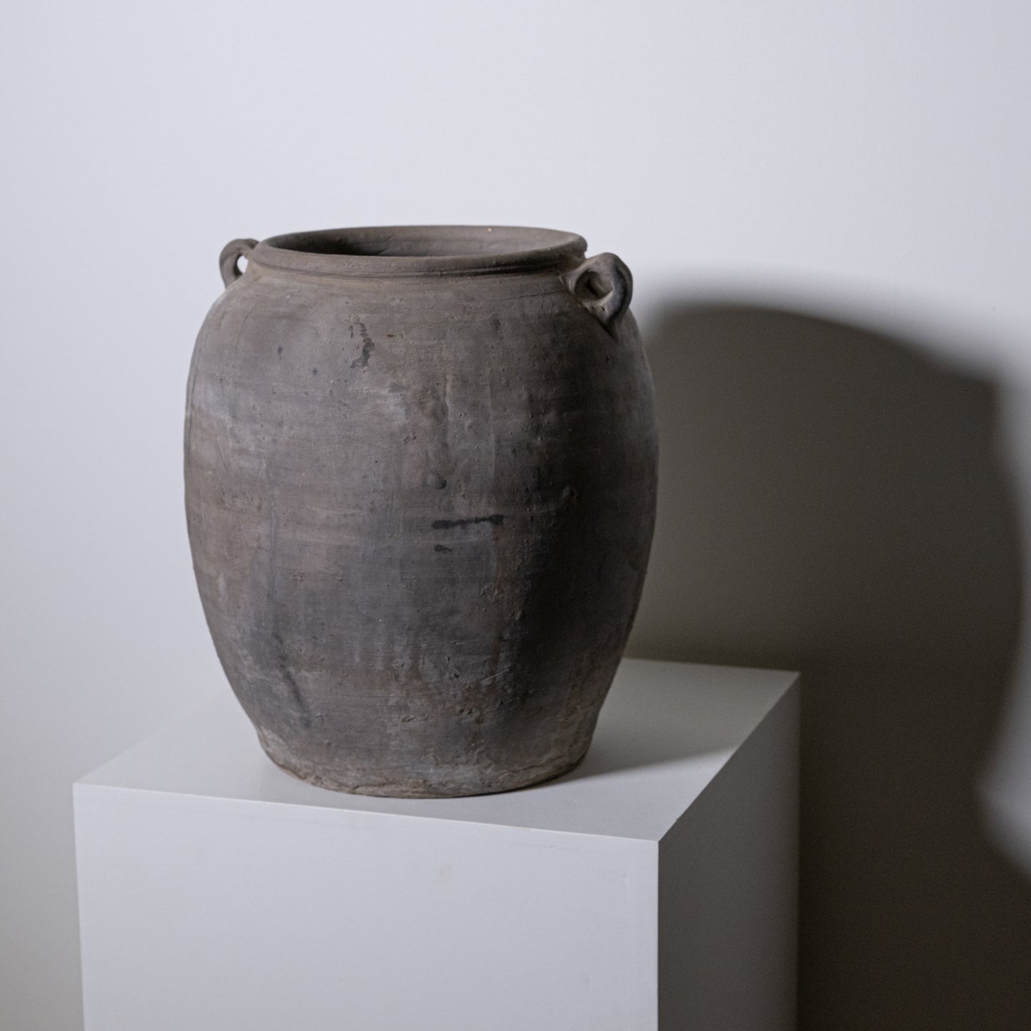 The 19th Century Central European Clay Pot, with its large round shape and two small handles by the rim, sits atop a white pedestal, adding antique charm to any decor with its dark clay finish and historic appeal.