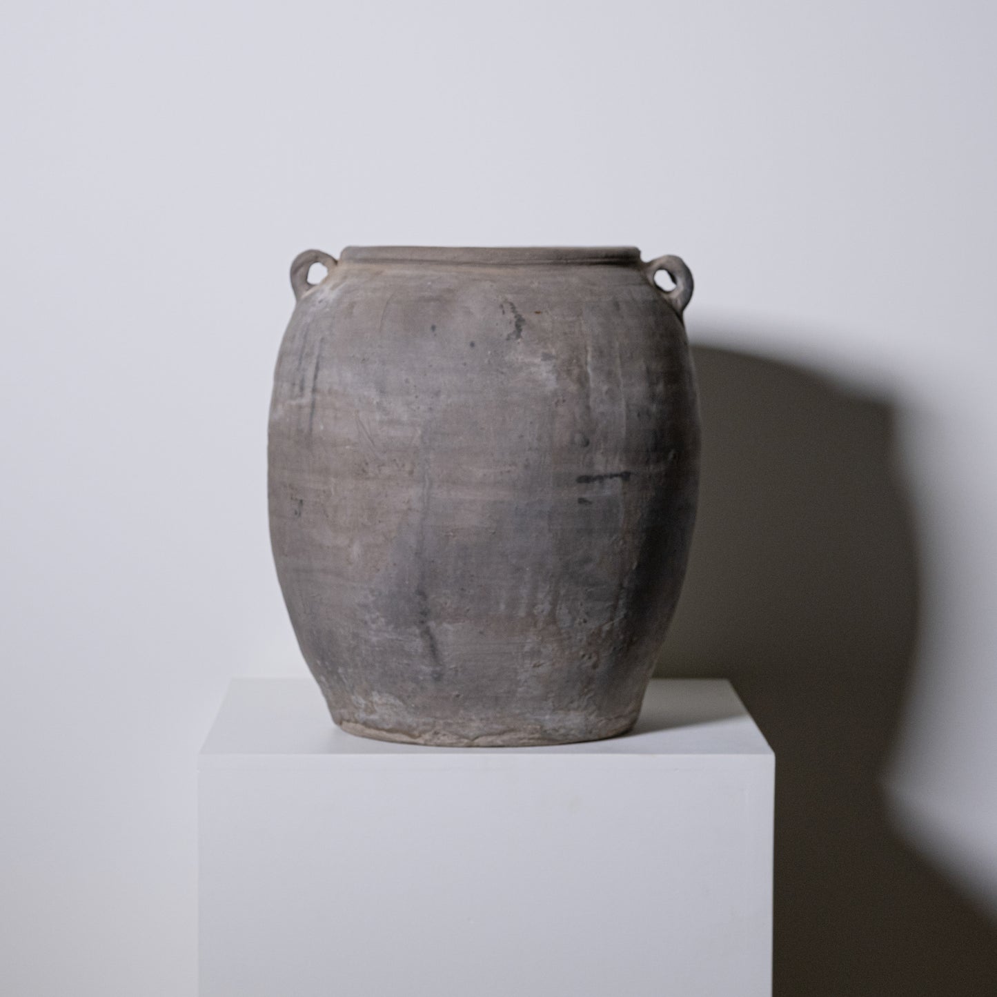 The 19th Century Central European Clay Pot is a large, weathered, round, grayish-brown jar with two small handles near the rim. Displayed on a white pedestal against a light background, it shows classic vintage wear and historic character.