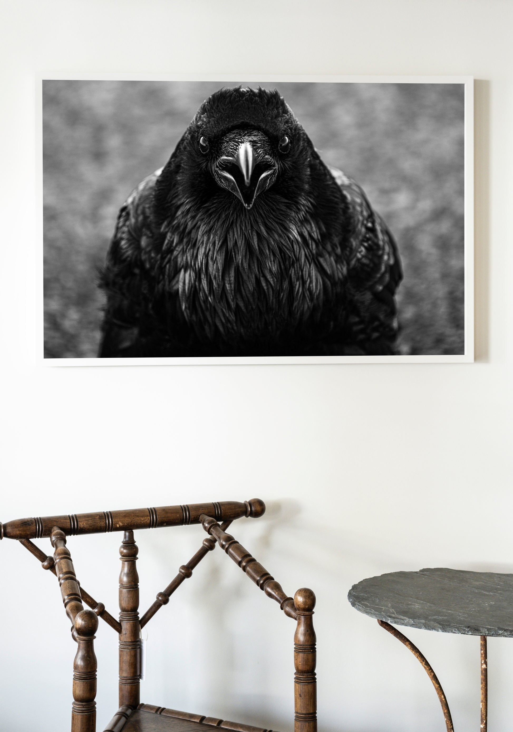 Framed black and white photograph of a raven on a wall above a wooden chair and small table.