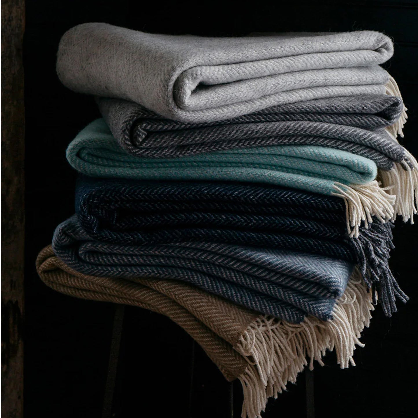 Stack of folded wool herringbone throw blankets in muted harvest tones with cream-colored fringe