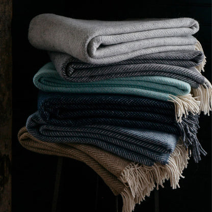 Stack of wool herringbone throw blankets in raw honey with cream-colored fringe and muted tones folded neatly