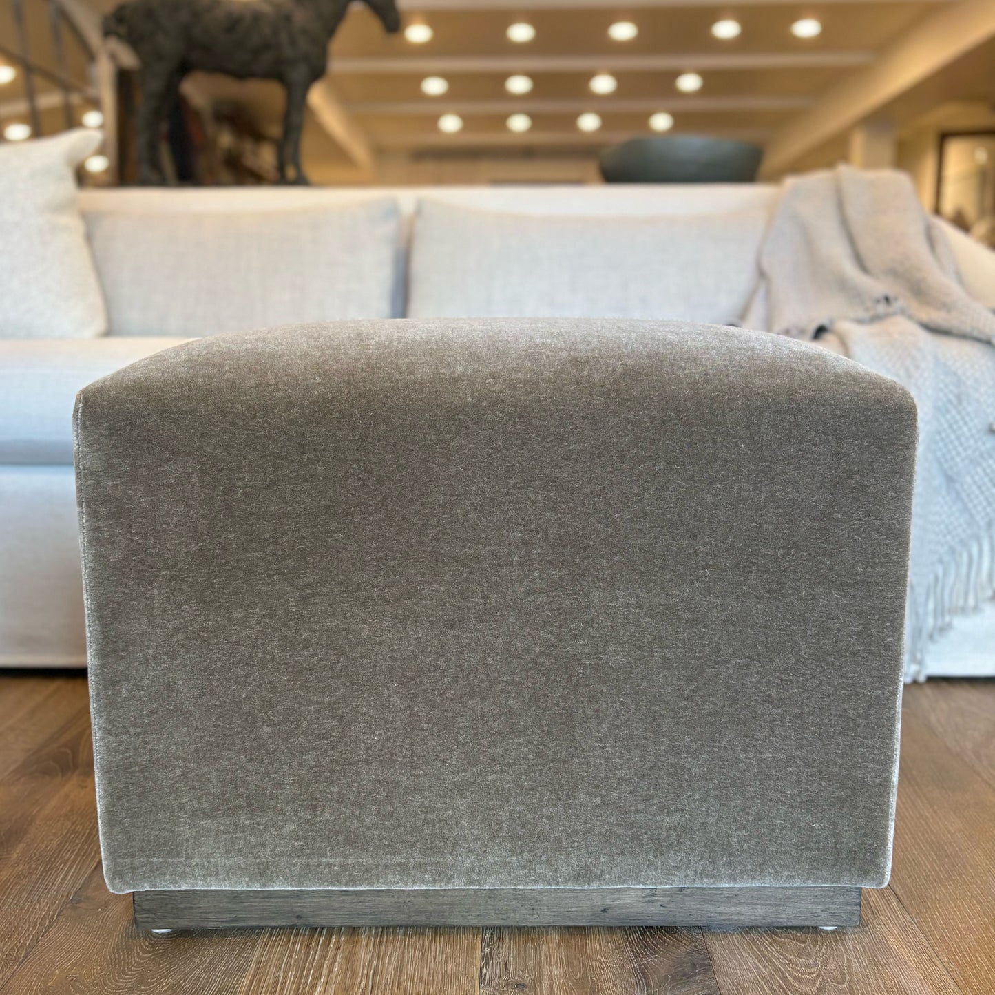 Gray upholstered aurora ottoman with textured fabric finish and dark wood base floor model