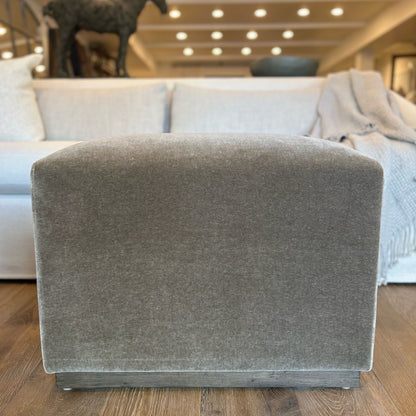 Gray upholstered aurora ottoman with textured fabric finish and dark wood base floor model