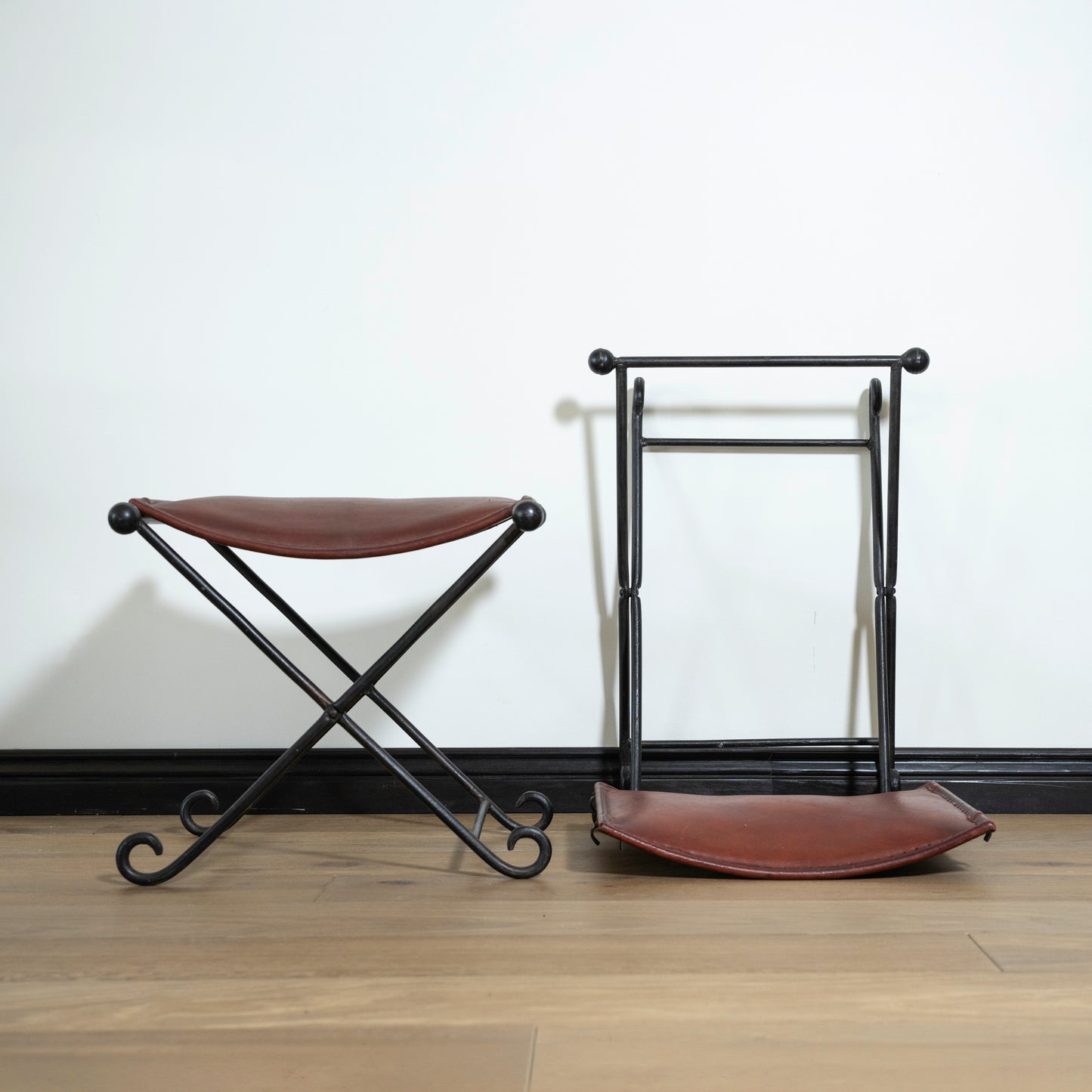 Two leather stools with metal frames on a wooden floor against a white wall.
