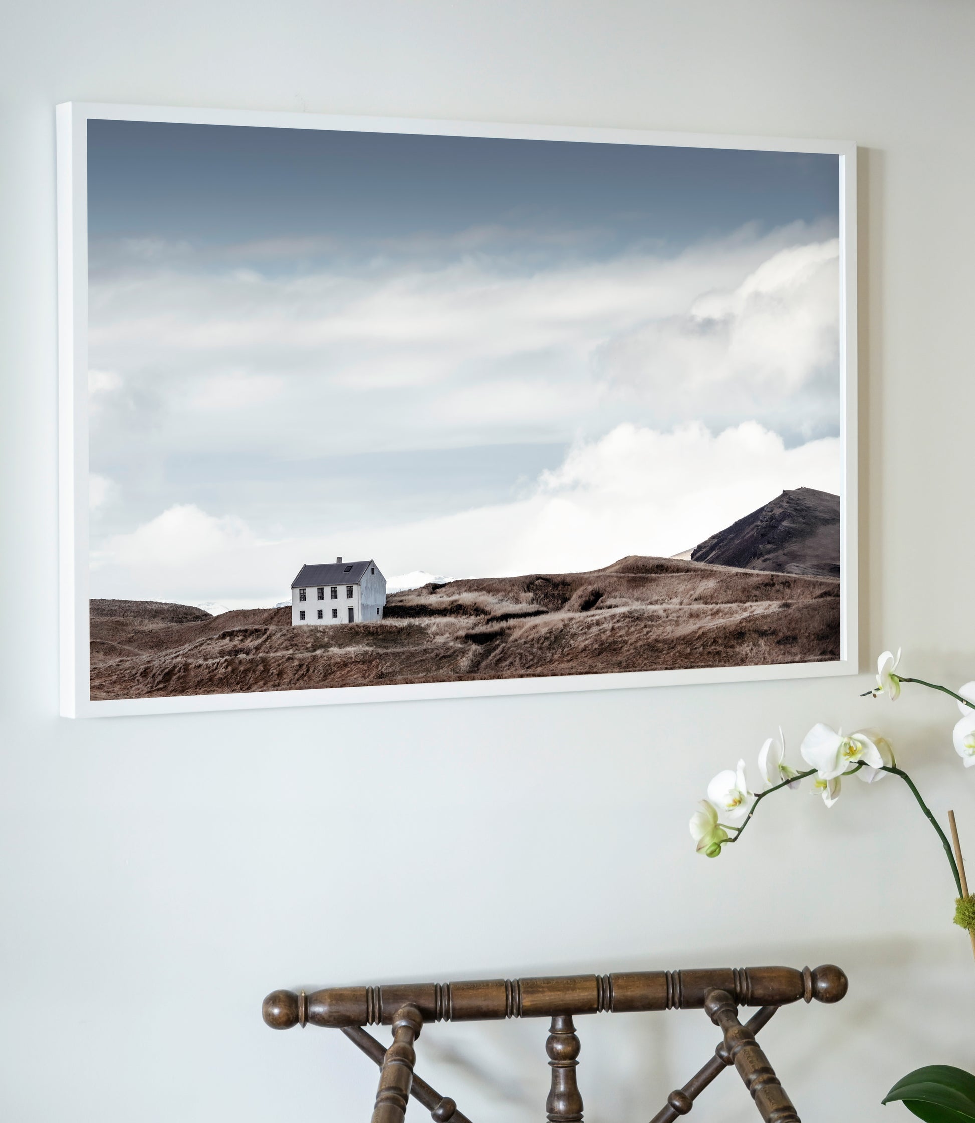 Framed landscape print of a house in a scenic setting on a wall above a wooden chair.