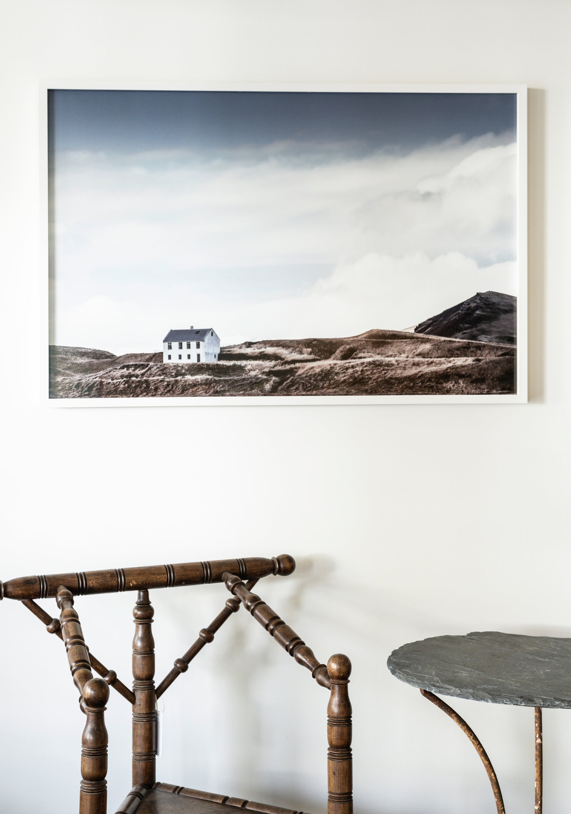 Framed landscape print of a house on a hillside above a wooden chair and table.