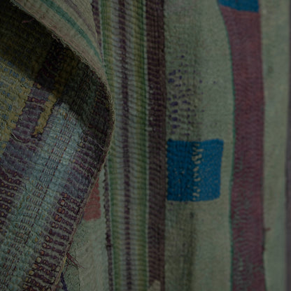 A close-up of a woven quilt with vertical stripes in muted green, purple, burgundy, and cream