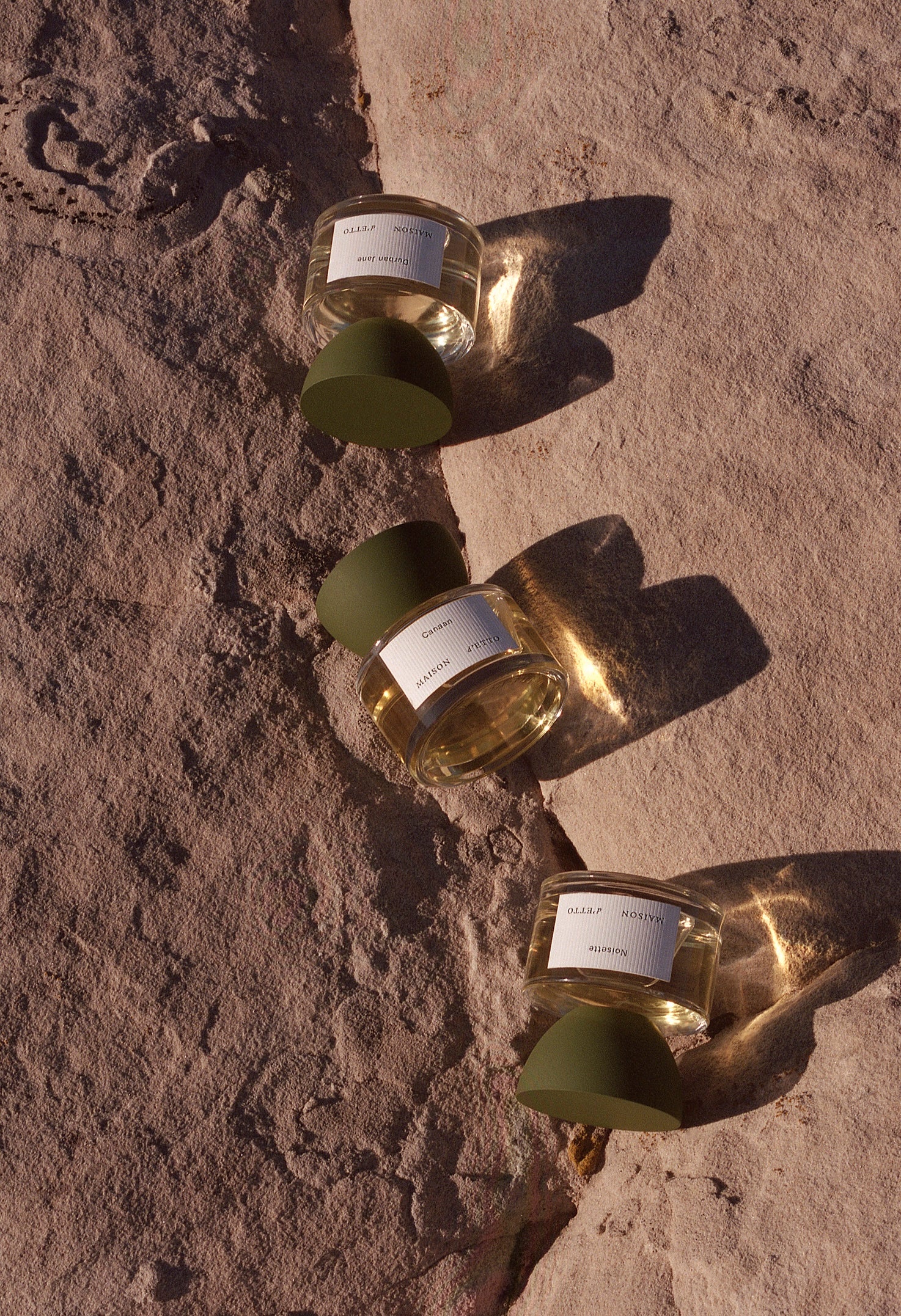 Three glass perfume bottles with olive green caps and minimalist white labels on textured stone surface for maison detto fragrance canaan