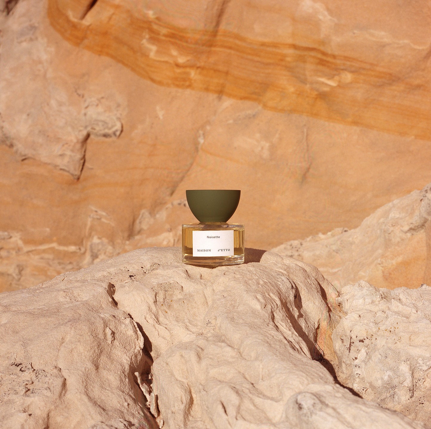 A minimalist glass perfume bottle with an olive green matte inverted bowl cap and mazette label set on sunlit desert rock