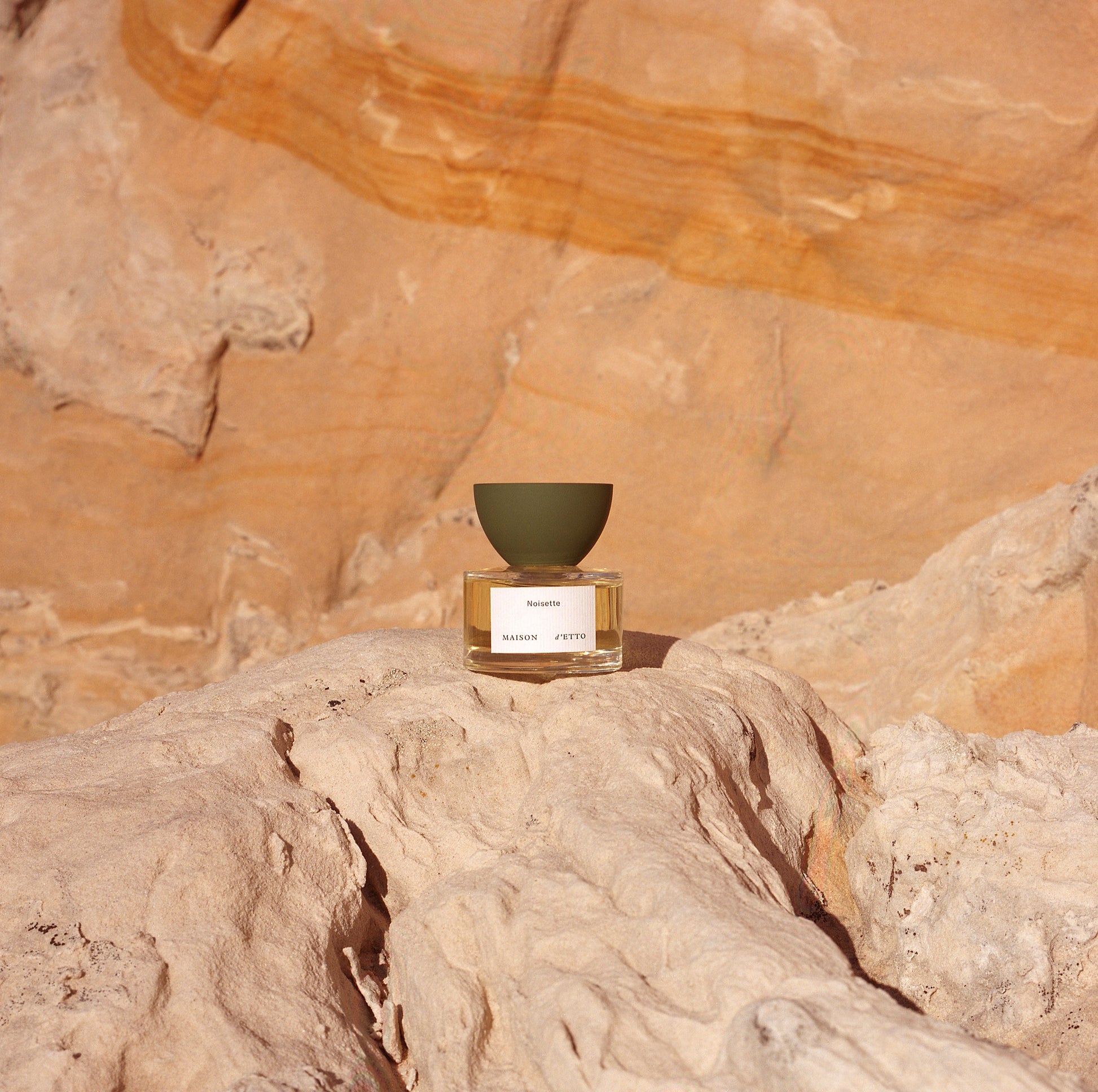 A minimalist glass perfume bottle with an olive green matte inverted bowl cap and mazette label set on sunlit desert rock