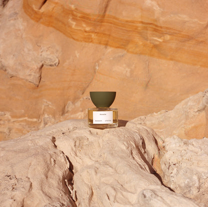 A minimalist glass perfume bottle with an olive green matte inverted bowl cap and mazette label set on sunlit desert rock
