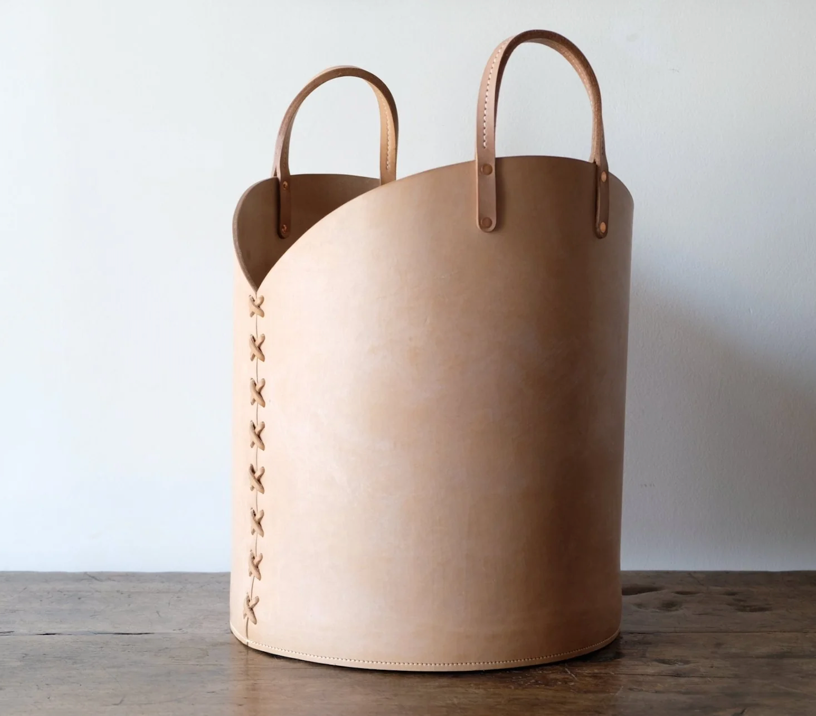 Tan leather log basket with curved handles and decorative x-stitching along the side for stylish storage