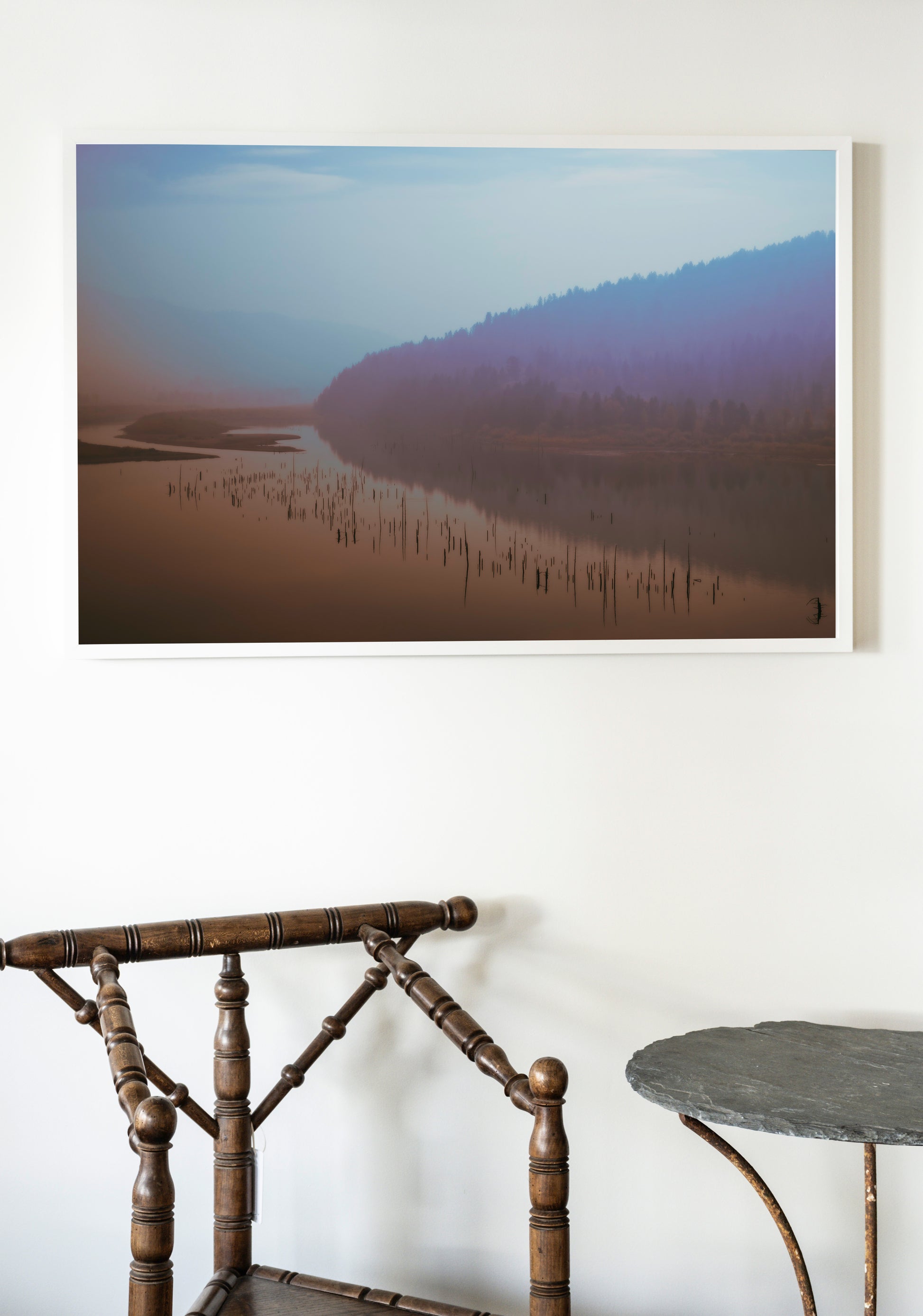 Framed photograph of a misty landscape with a lake and mountains on a white wall above a wooden chair and table.