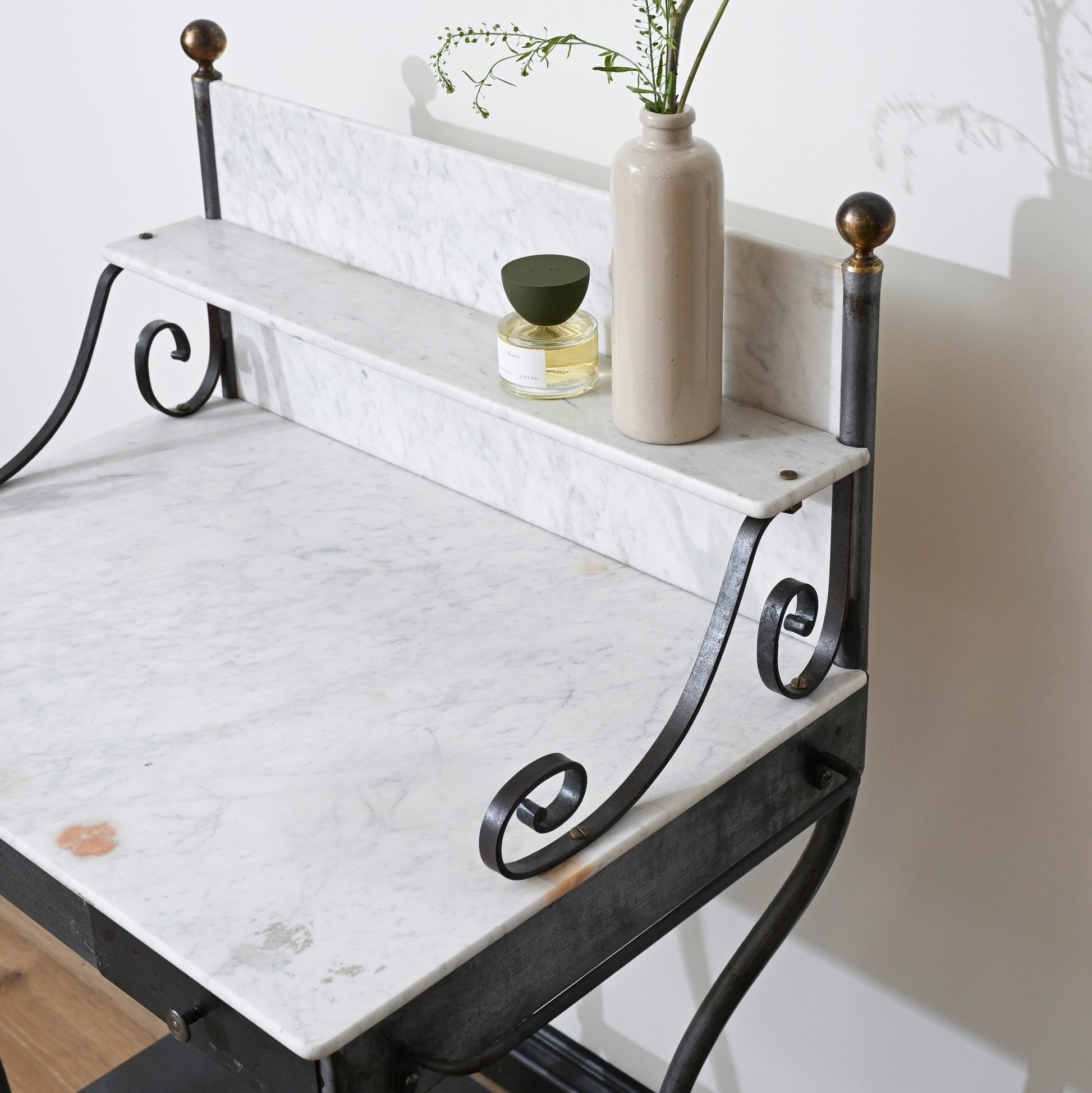 A marble-top desk with black metal legs and ornate scroll details, featuring a raised marble shelf holding a beige vase with greenery, a small glass jar, and the Antique Shanxi Ceramic Vessels (Set of 3), is set against a white wall.
