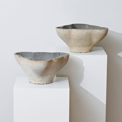 Two textured, handmade ceramic bowls with uneven edges sit on white pedestals against a white background. Inspired by the 19th Century Antique Belgian Trefoil Planter, they are light beige with gray interiors.