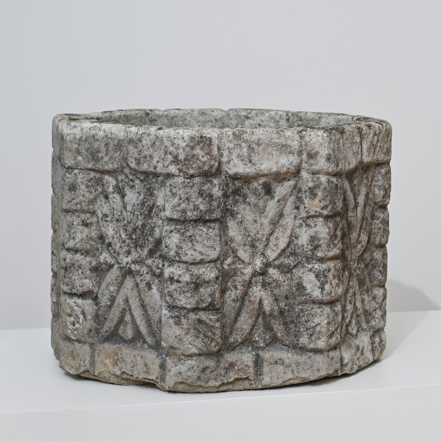The 19th Century English Concrete Jardinere is a cylindrical, weathered stone planter featuring rough surfaces with repeating geometric and floral carvings, shown against a plain white background.