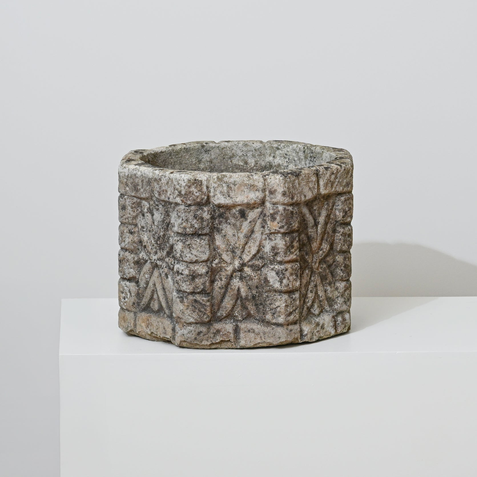 The 19th Century English Concrete Jardinere, featuring a round, carved form with rough texture and geometric patterns, sits on a white pedestal against a light gray background, evoking timeless English elegance.