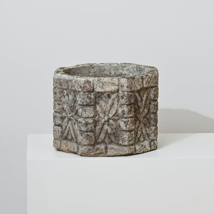 The 19th Century English Concrete Jardinere, featuring a round, carved form with rough texture and geometric patterns, sits on a white pedestal against a light gray background, evoking timeless English elegance.
