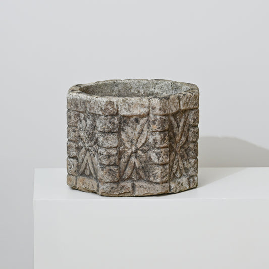 The 19th Century English Concrete Jardinere, featuring a round, carved form with rough texture and geometric patterns, sits on a white pedestal against a light gray background, evoking timeless English elegance.