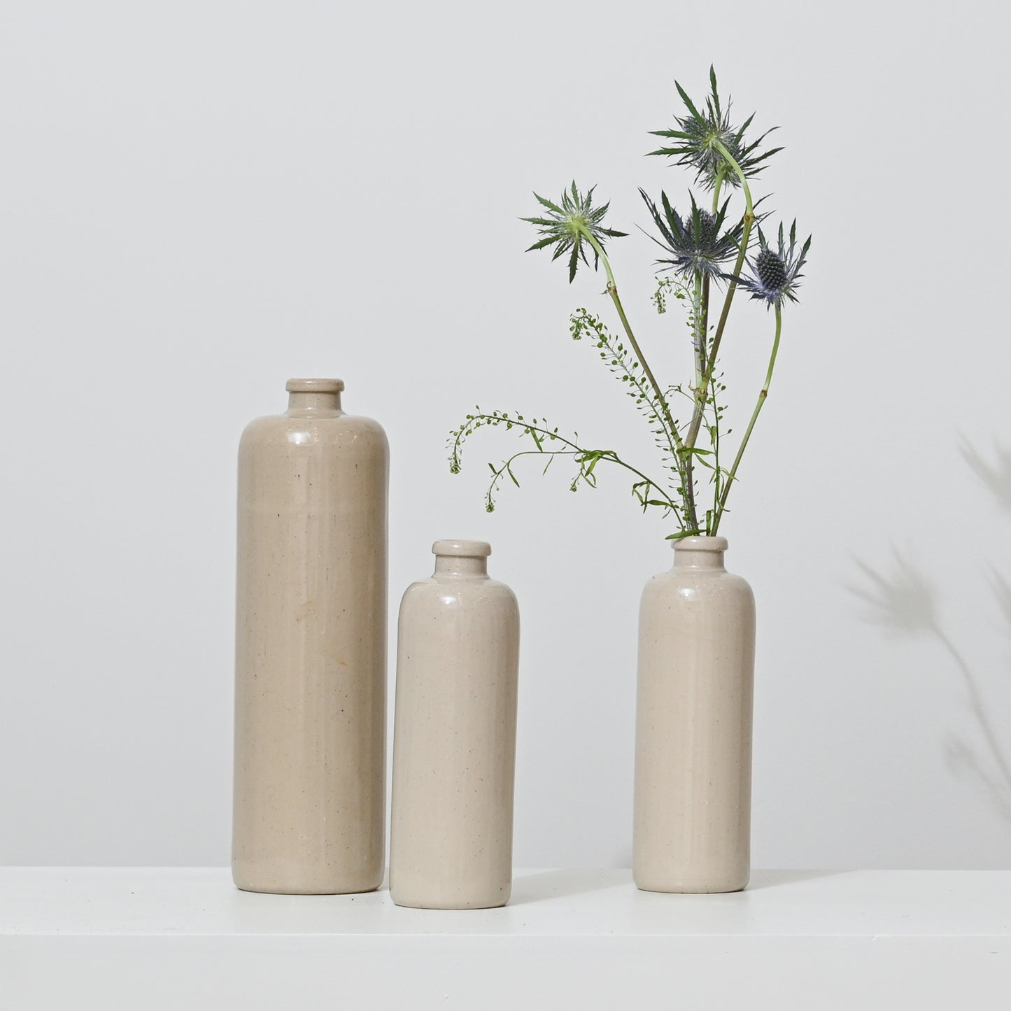 The Antique Shanxi Ceramic Vessels (Set of 3)—beige, varying in height, and inspired by antique Chinese jars—are arranged on a white surface; the tallest holds green and spiky purple thistle flowers while the others are empty.