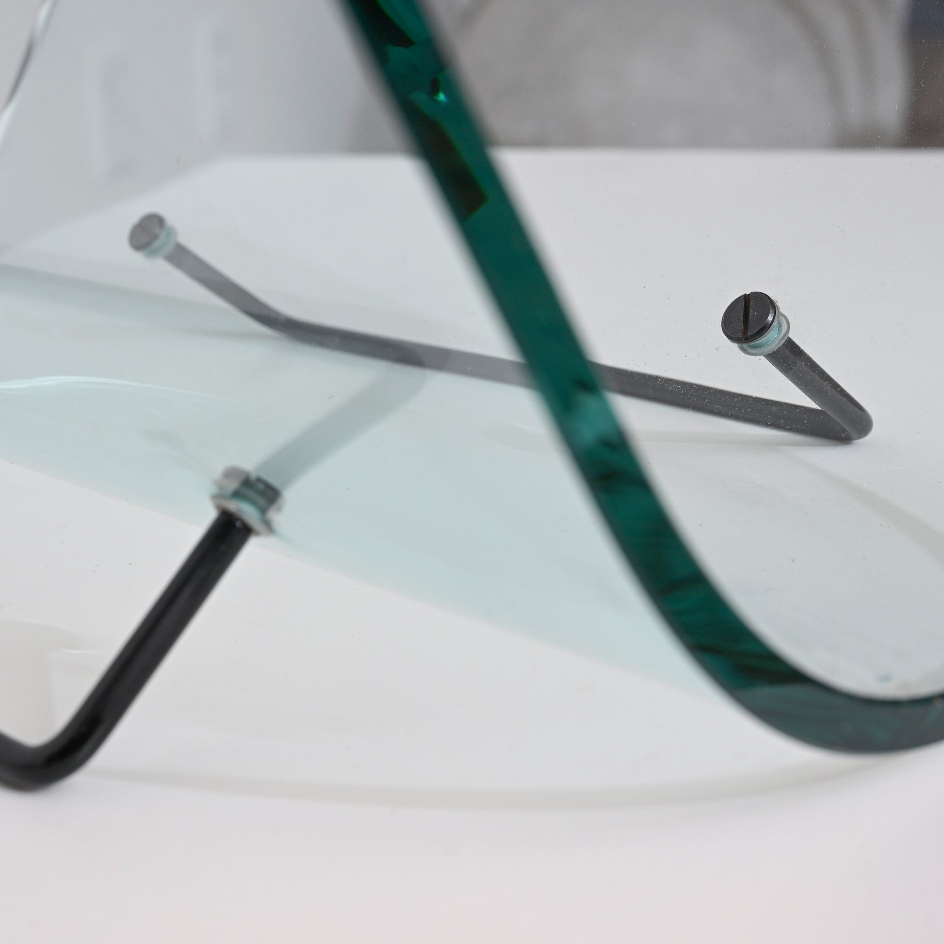 A close-up of the Vintage Italian Curved Glass Magazine Holder shows its curved glass sheet secured by black metal rods with screws, highlighting its modern Italian design on a white surface.