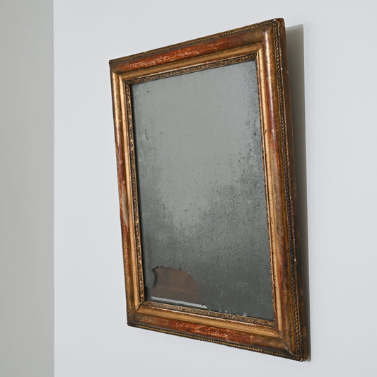 An Early 19th Century Mercury Glass Mirror with a rectangular, distressed gilt wood frame hangs on a white wall; its heavily tarnished surface shows dark spots and significant damage near the bottom edge.