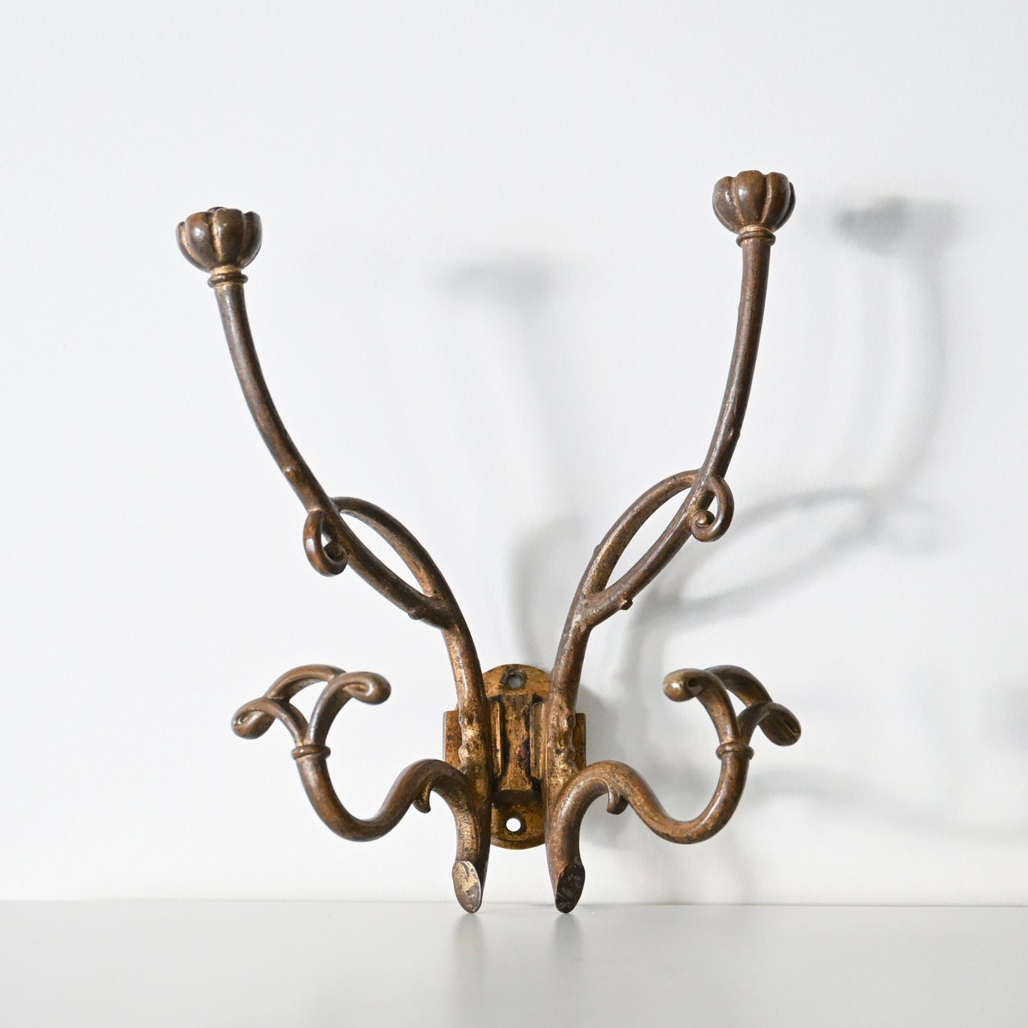 The Antique French Bronze Hook features four curved arms with floral-shaped ends, inspired by 1930s French decor. Its ornate, wall-mounted design casts elegant shadows on a white background.
