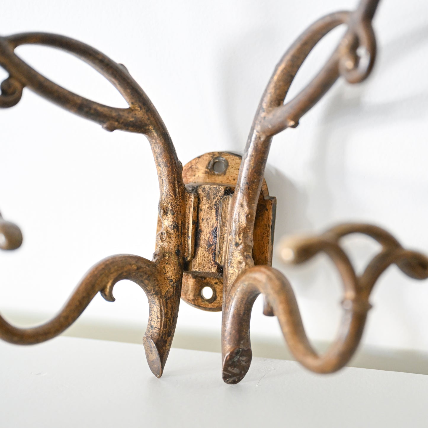 A close-up of the Antique French Bronze Hook with curved decorative arms and an aged rustic finish, inspired by 1930s French decor, mounted on a white wall.