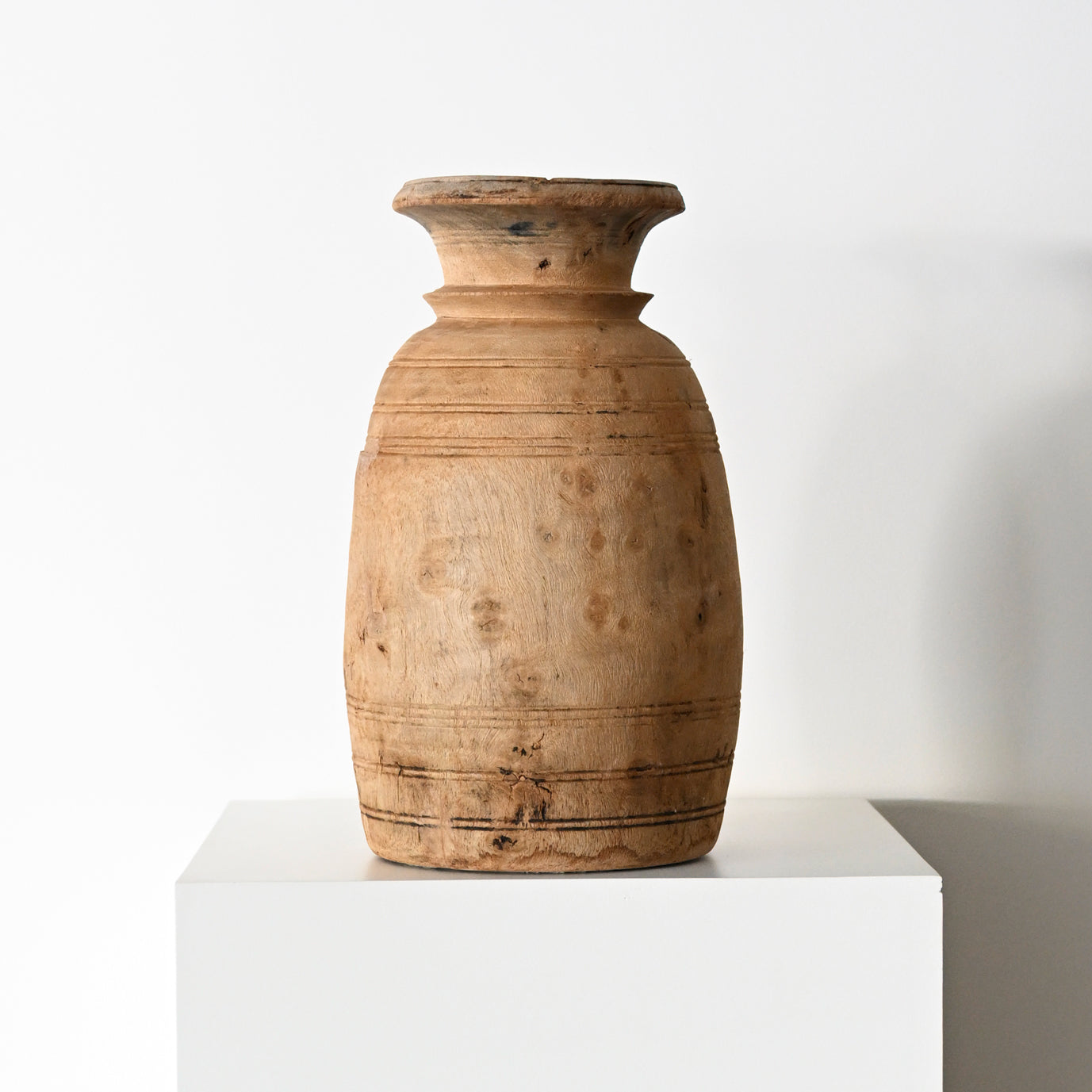 A rustic light brown wooden vase with subtle horizontal carvings and natural wood grain from primitive wood vessels