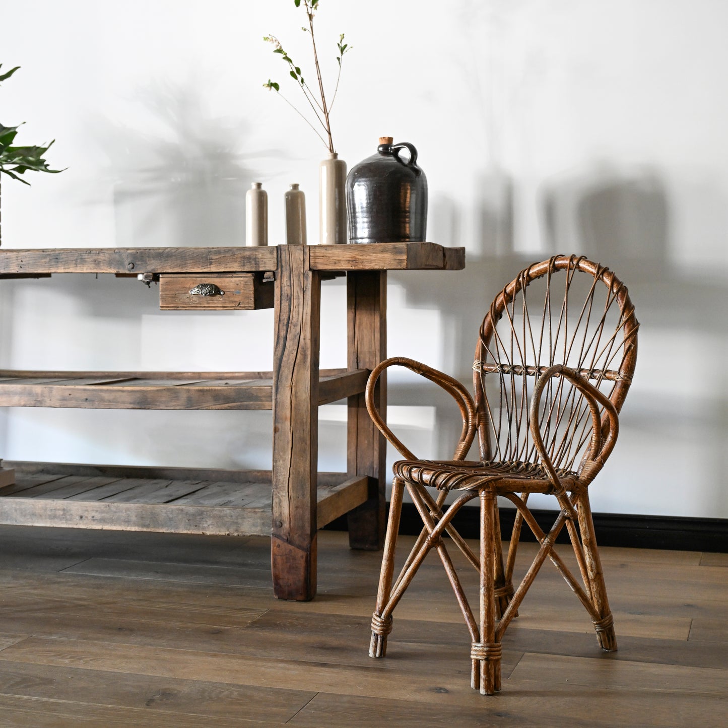 A rustic wooden table with shelves stands next to a rattan chair on a wooden floor. On the table are three Antique Shanxi Ceramic Vessels, a black jug, and other antiques, with branches arranged in one vessel.