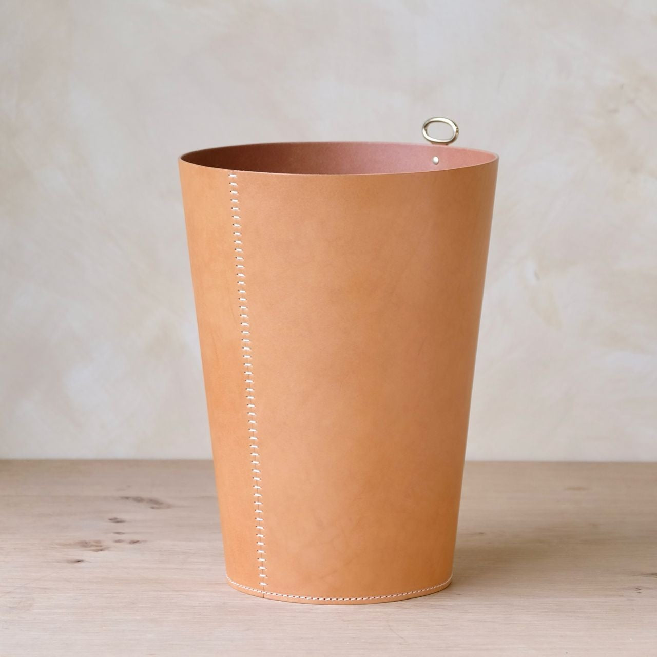 Eyelet leather wastepaper bin in tan leather with white stitching and a silver metal handle for desk use