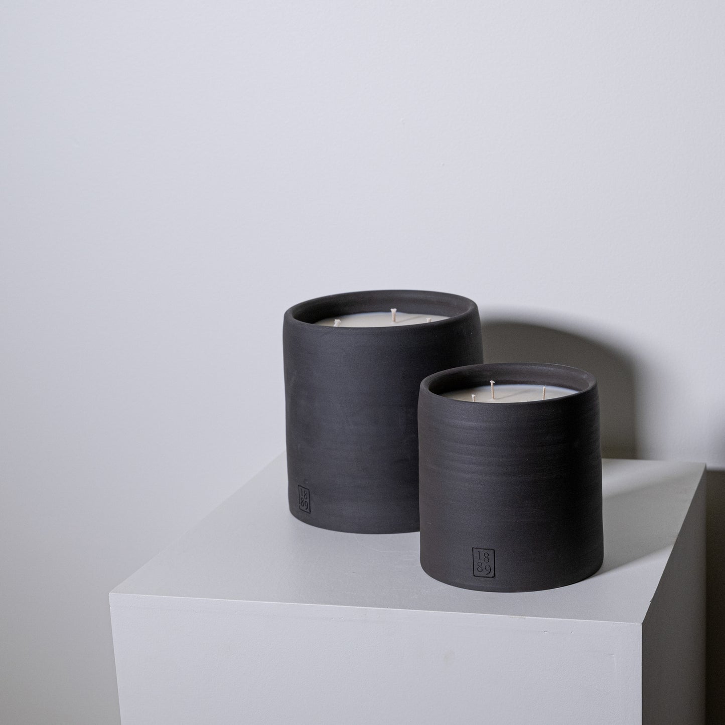Two matte black cylindrical candles with white wax and multiple wicks from candle savu with embossed logos on base