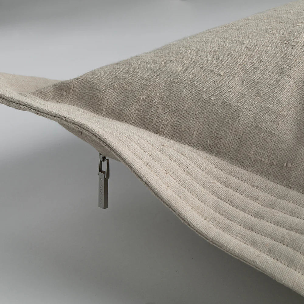 Beige linen bucket hat with ribbed brim and metallic zipper pull shown next to quilted edge pillow cement color