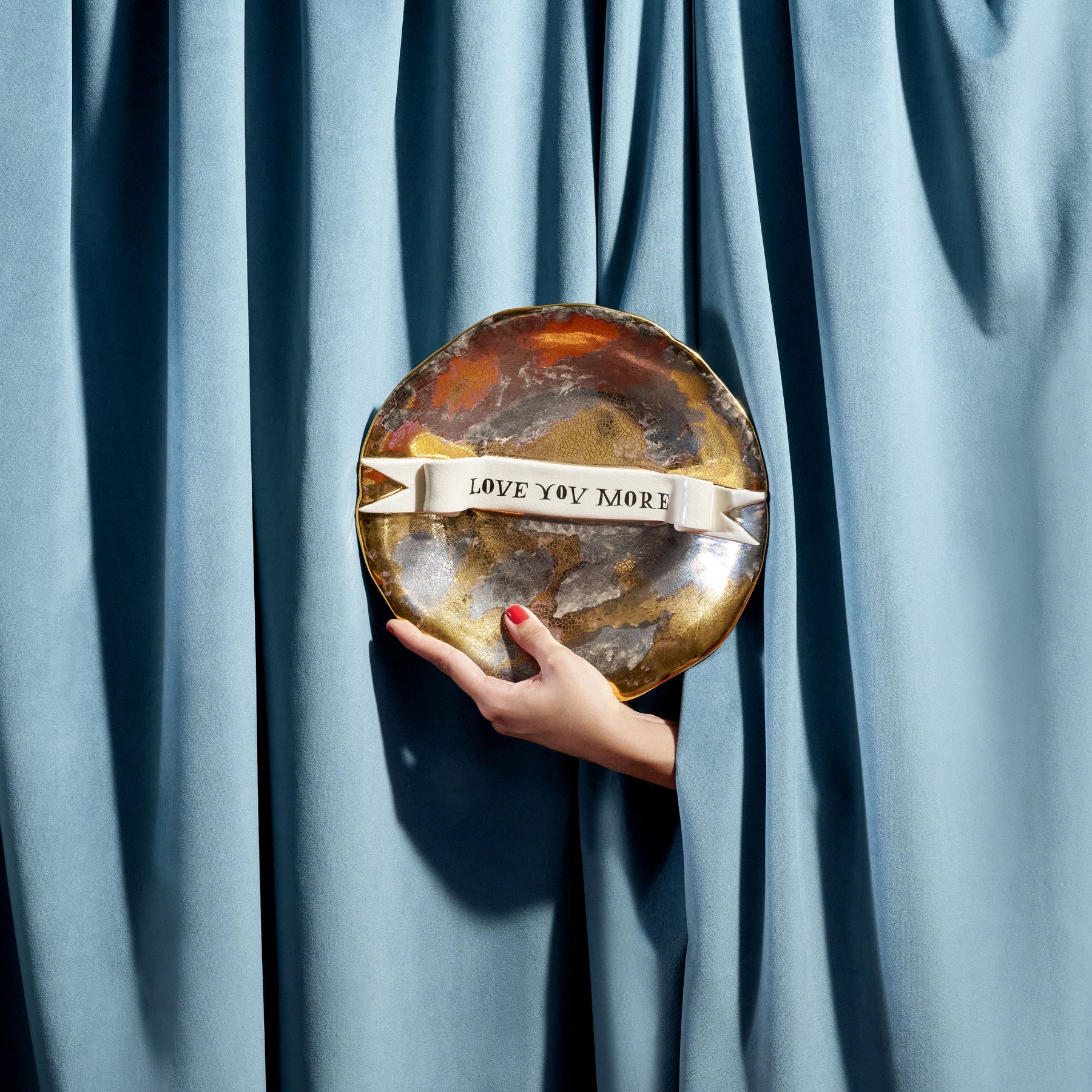 A hand holds a round metallic Ruan Hoffman love you more plate with marbled gold and silver finish and white ribbon