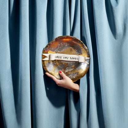A hand holds a round metallic Ruan Hoffman love you more plate with marbled gold and silver finish and white ribbon