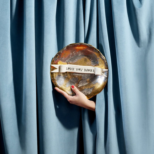 A hand holds a round metallic Ruan Hoffman love you more plate with marbled gold and silver finish and white ribbon